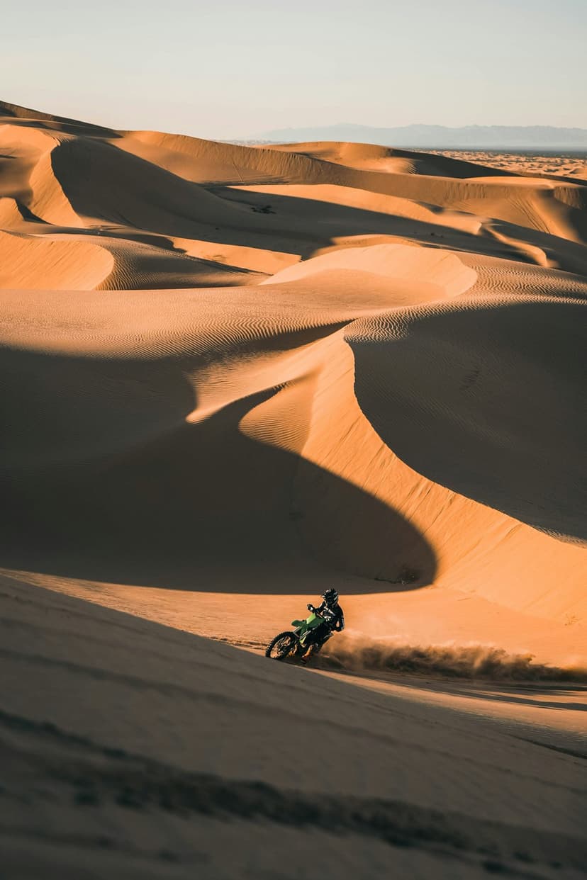 Motocross rider on a green dirt bike kicks up sand while ascending a steep dune under a glowing golden sunset.