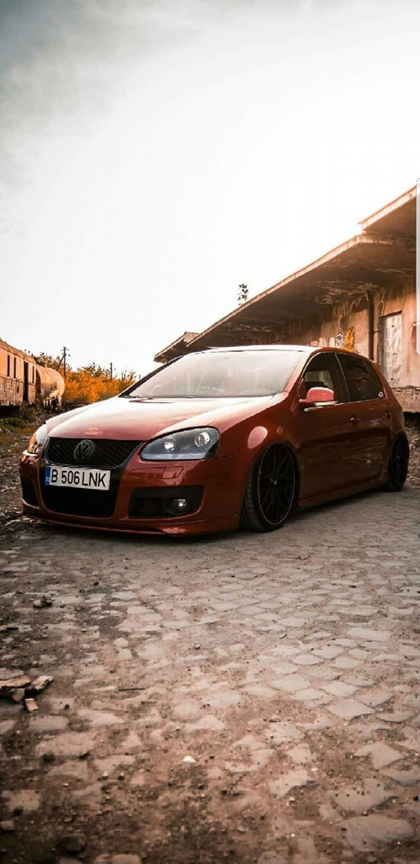 Red Volkswagen Golf GTI with lowered suspension parked on a grey cobblestone street beside an industrial brick wall.