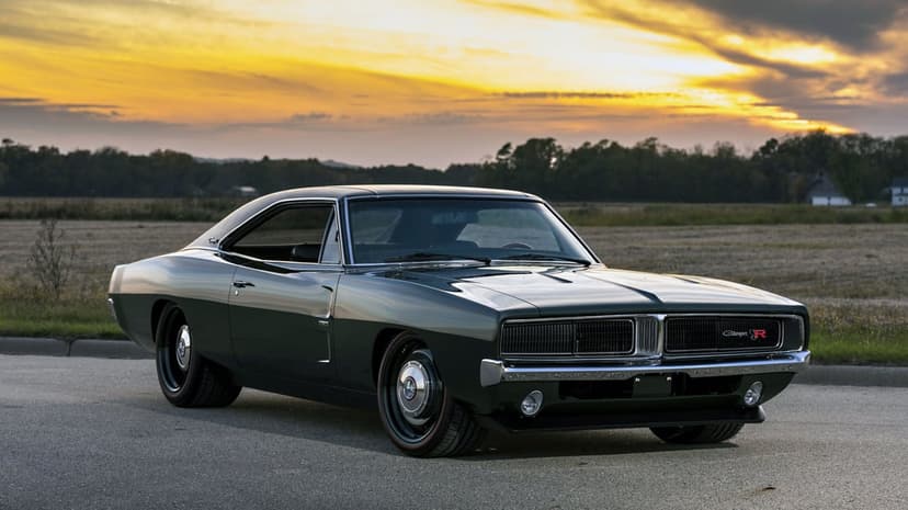 Dark green 1969 Dodge Charger R/T parked on asphalt against a vibrant orange sunset sky and rural landscape.