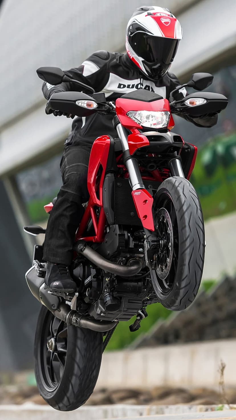 Ducati rider in black and white leathers pops a wheelie on a red sportbike against a blurred urban background.