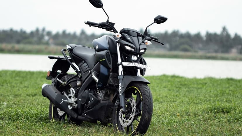 Yamaha MT-15 motorcycle in dark matte blue parked on green grass with a blurred lake and forest background.