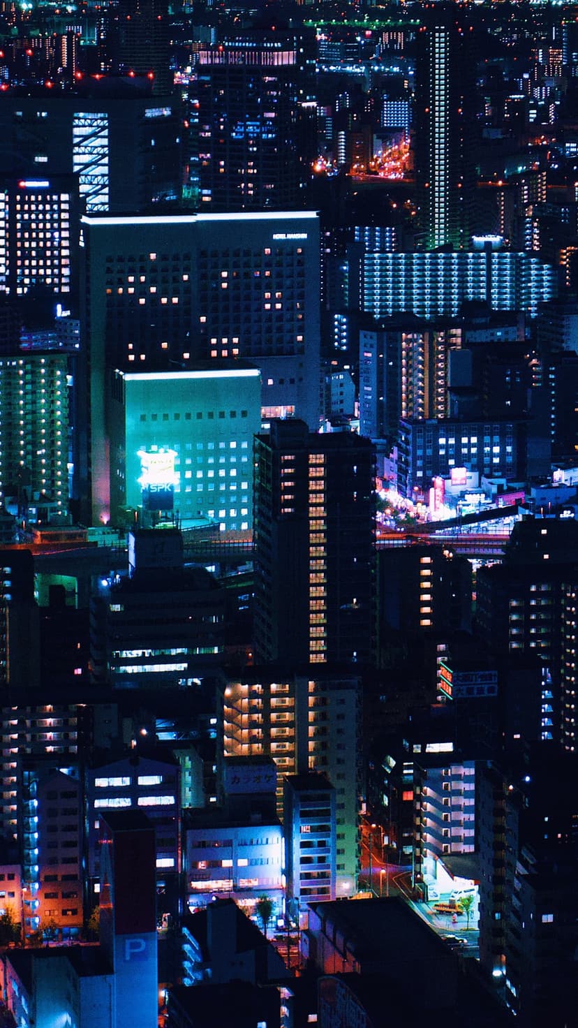 Illuminated Tokyo skyscrapers glow with blue and amber lights across a dense night skyline in a vertical view.