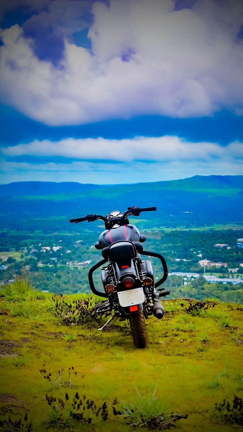 Classic black motorcycle parked on a grassy ridge overlooking a vast green valley under a cloudy blue sky.