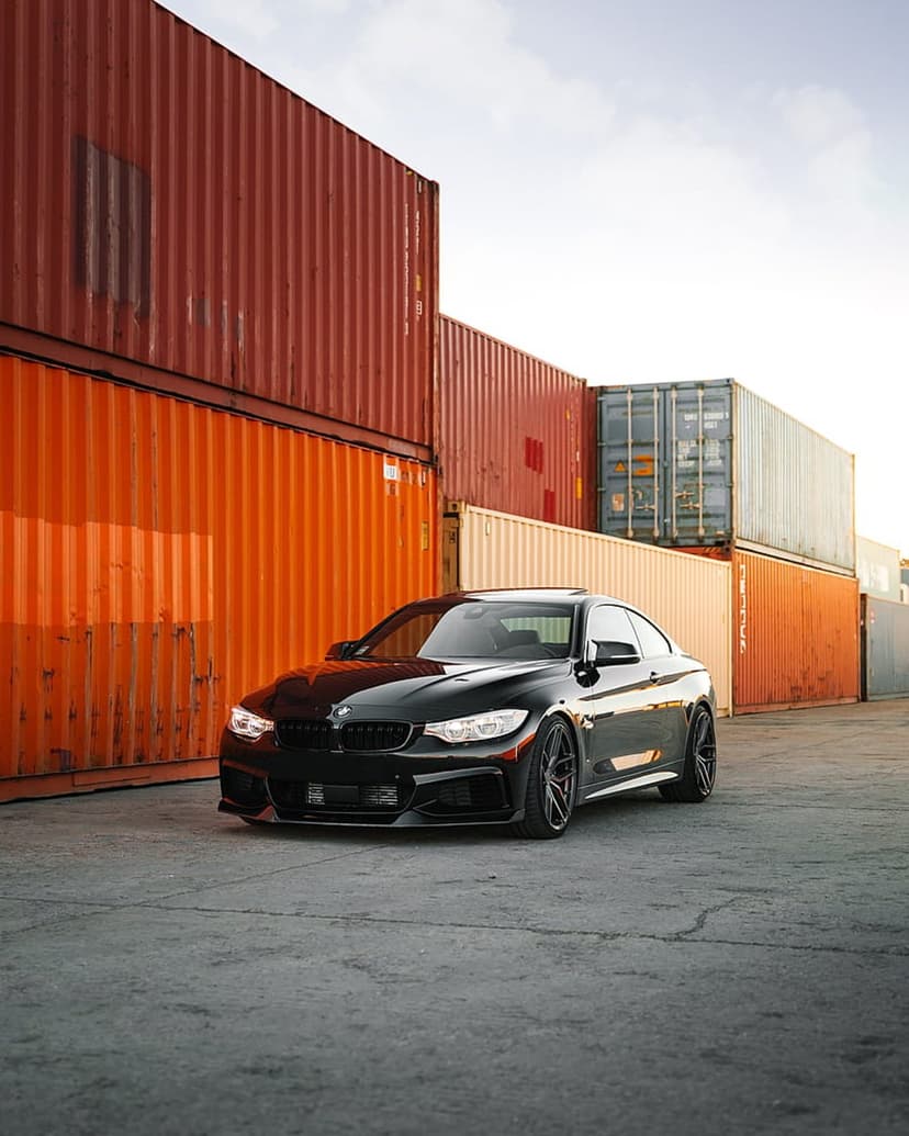 Black BMW M4 coupe parked beside stacked orange shipping containers under a soft blue and white sunset sky.