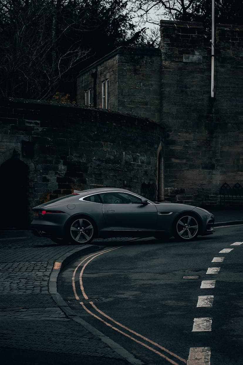 Dark gray Jaguar F-Type coupe parked on a winding cobblestone street next to an old stone building in moody light.