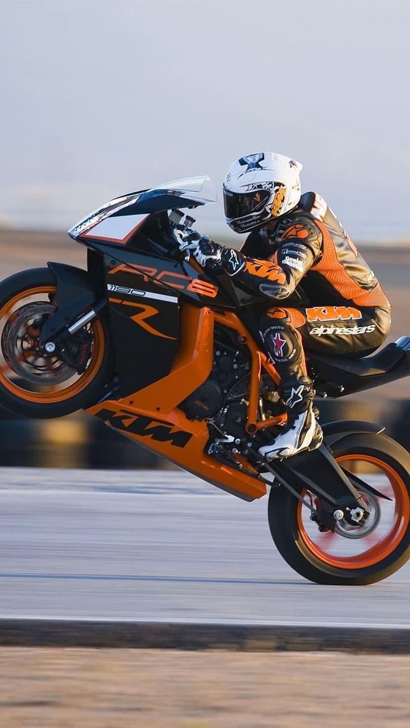 Motorcyclist in an orange racing suit performs a wheelie on a KTM sportbike across an asphalt track under a blue sky.