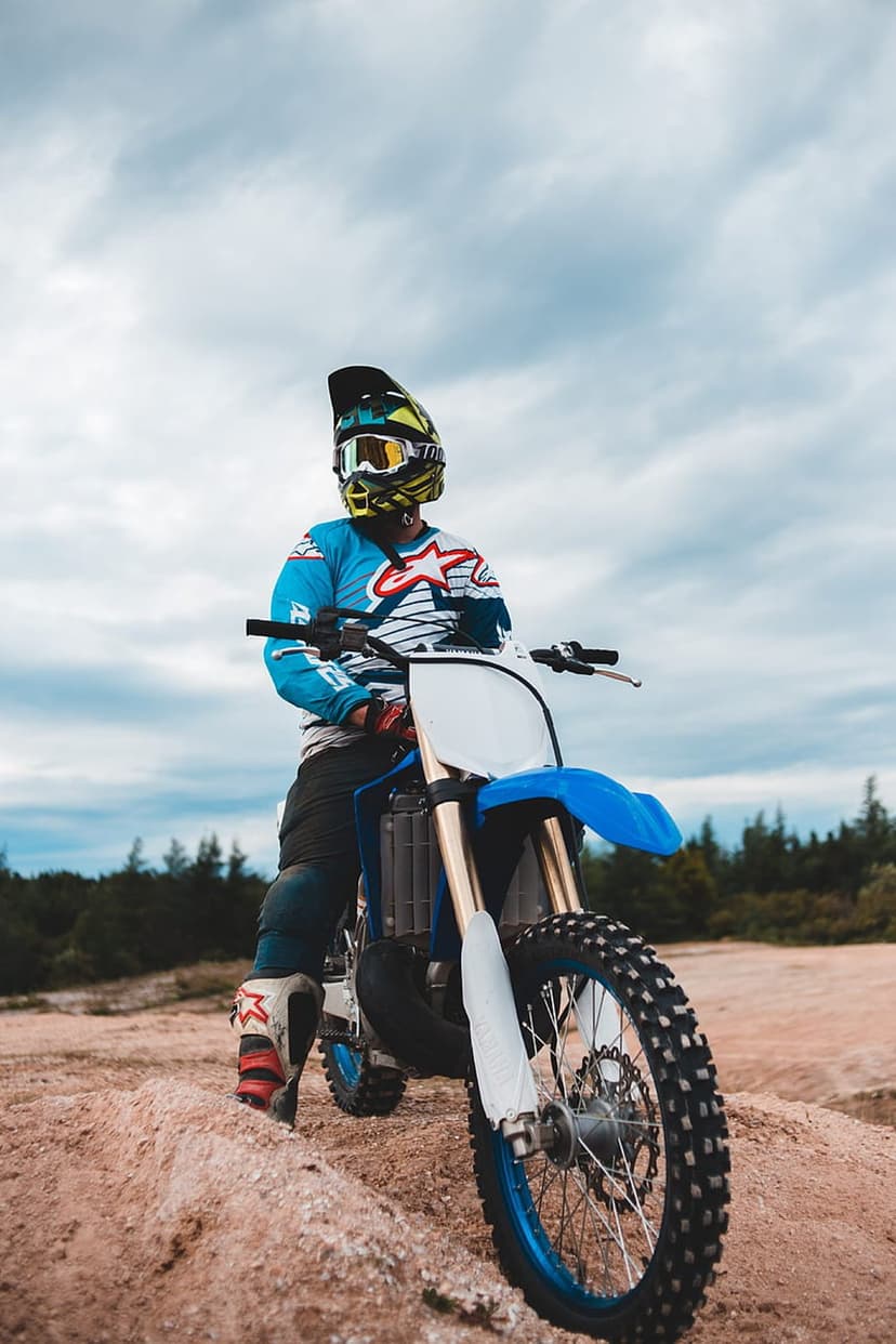 Motocross rider in blue and white racing gear sits on a dirt bike on a sandy outdoor track under a cloudy sky.