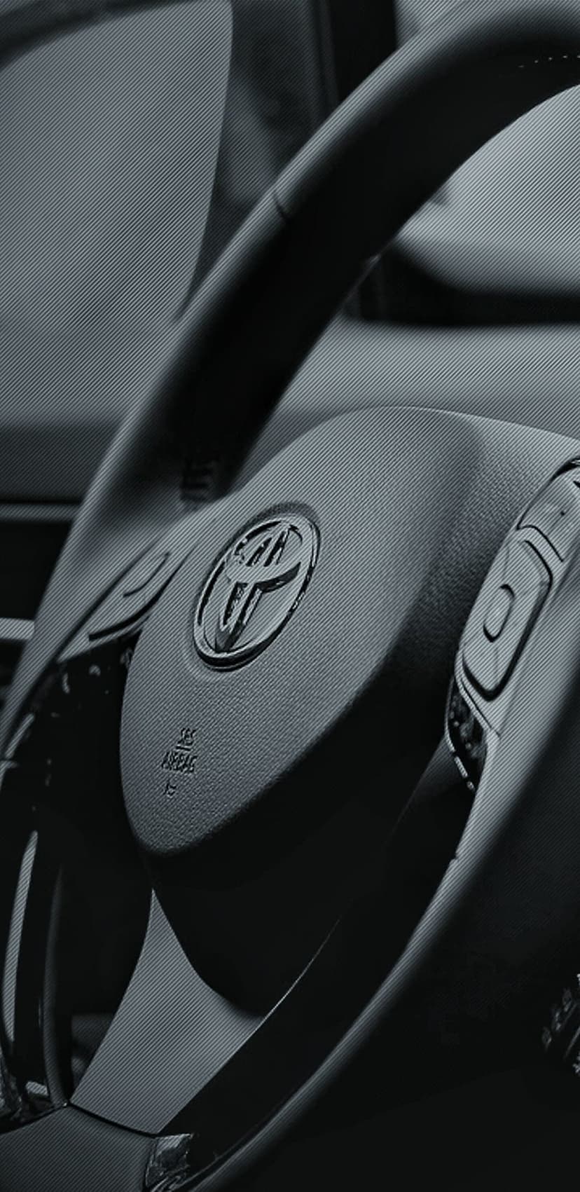 Toyota steering wheel with a silver emblem and side control buttons in a textured dark automotive interior.