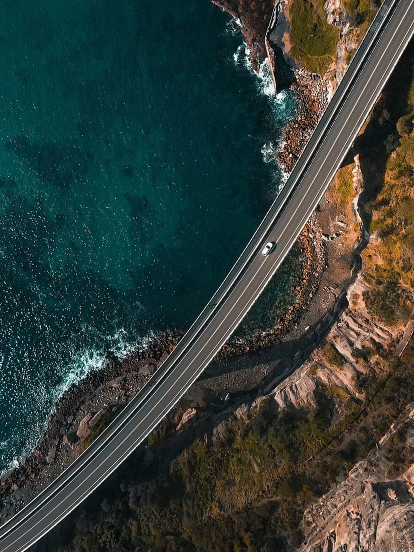White car drives across a curved bridge overlooking a teal ocean and rocky coastline with green vegetation.