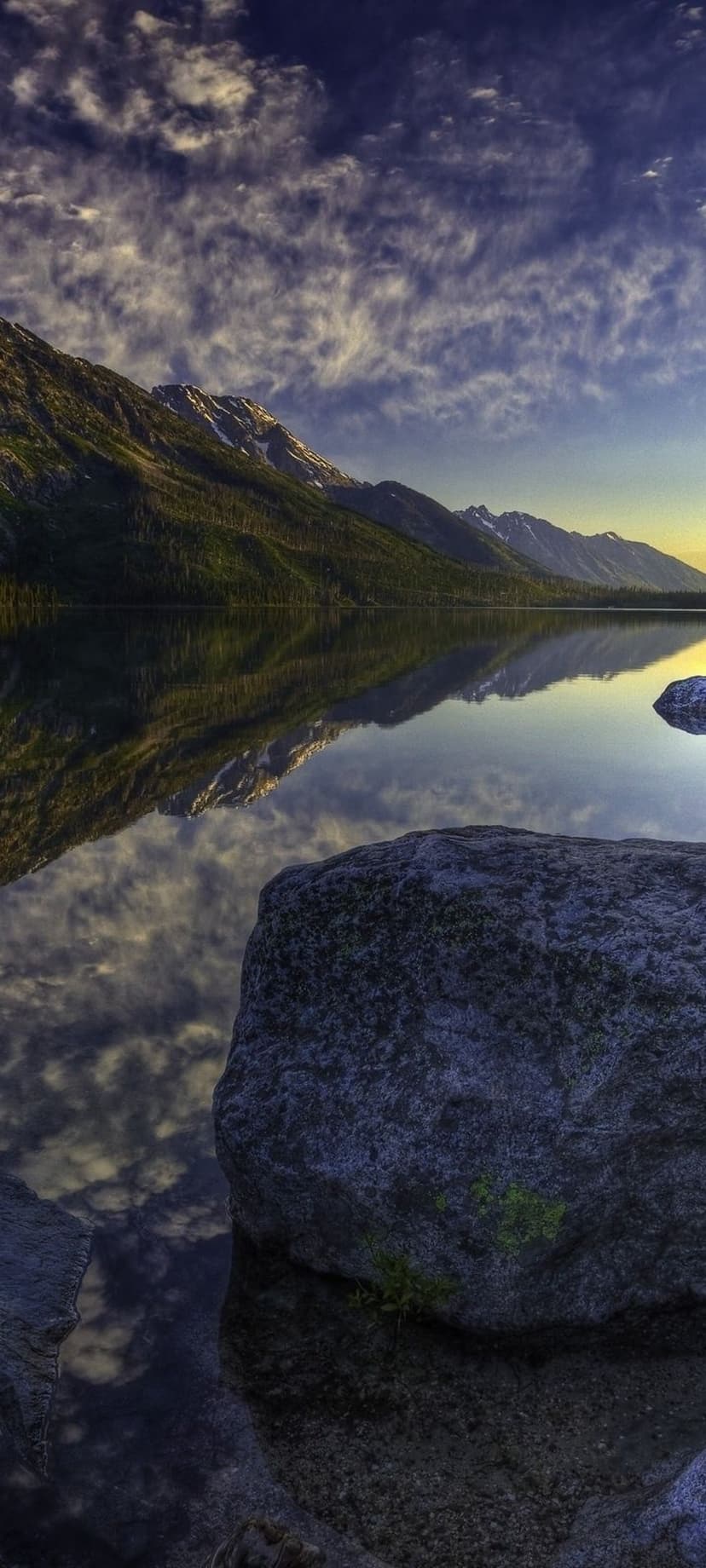 Sharp mountain peaks reflect in a calm glacial lake surrounded by large rocks under a colorful sunset sky.