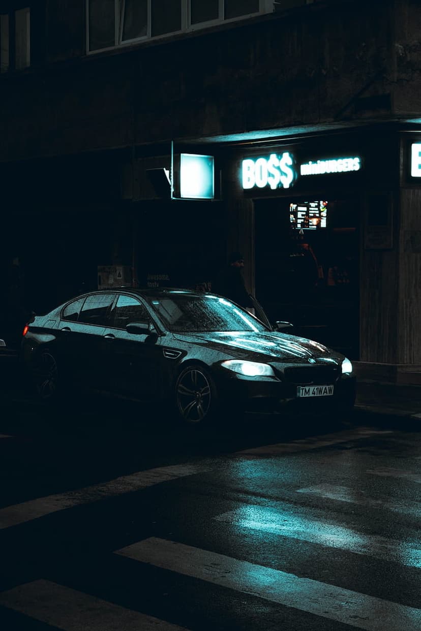 Black BMW sedan parked at a wet crosswalk illuminated by a glowing neon burger sign and bright headlights.