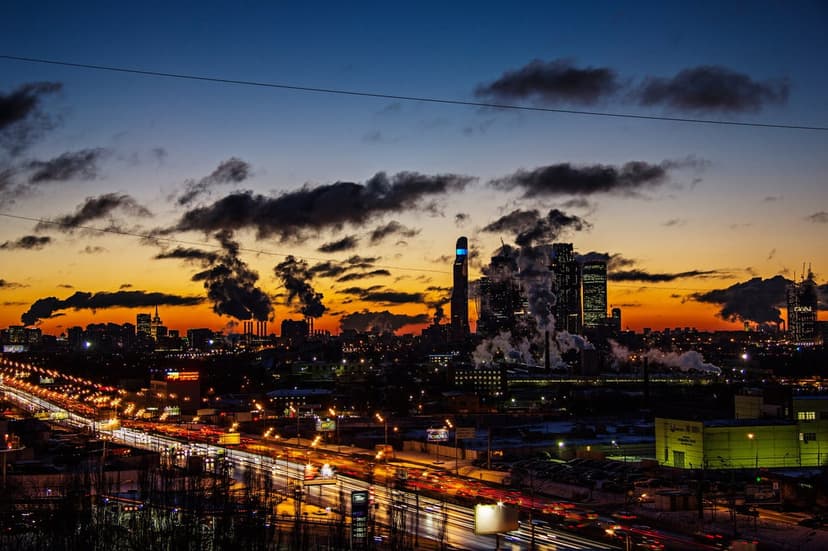 Towering factory chimneys release smoke plumes against a vivid orange and blue sunset sky above a glowing highway.