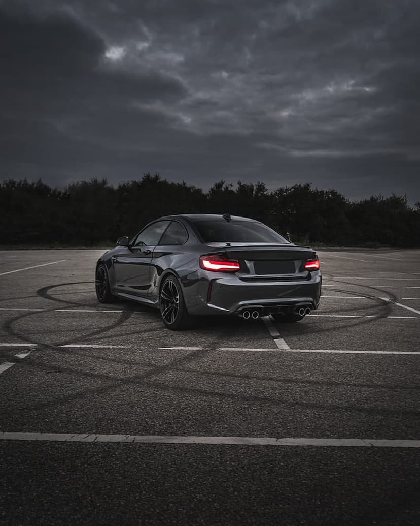 BMW M2 coupe with glowing red taillights and tire marks on asphalt under a dark, dramatic stormy sky.