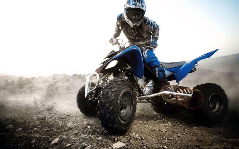 Rider on a blue Yamaha ATV kicking up thick dust on a rocky dirt path under a bright, high-key sunlit sky.