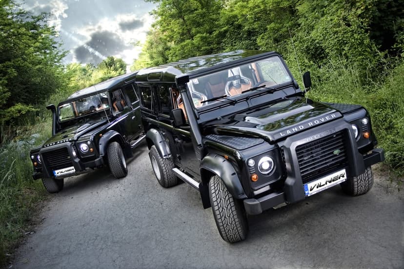 Two black Land Rover Defenders parked on a gravel path surrounded by dense green forest under a cloudy sky.