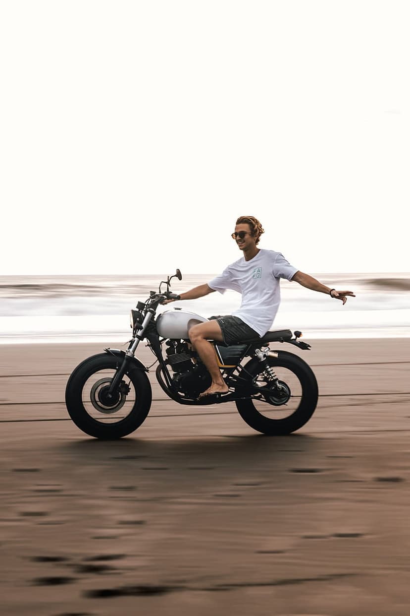 Blonde man riding a white motorcycle on a sandy beach with arms spread wide against a backdrop of ocean waves.