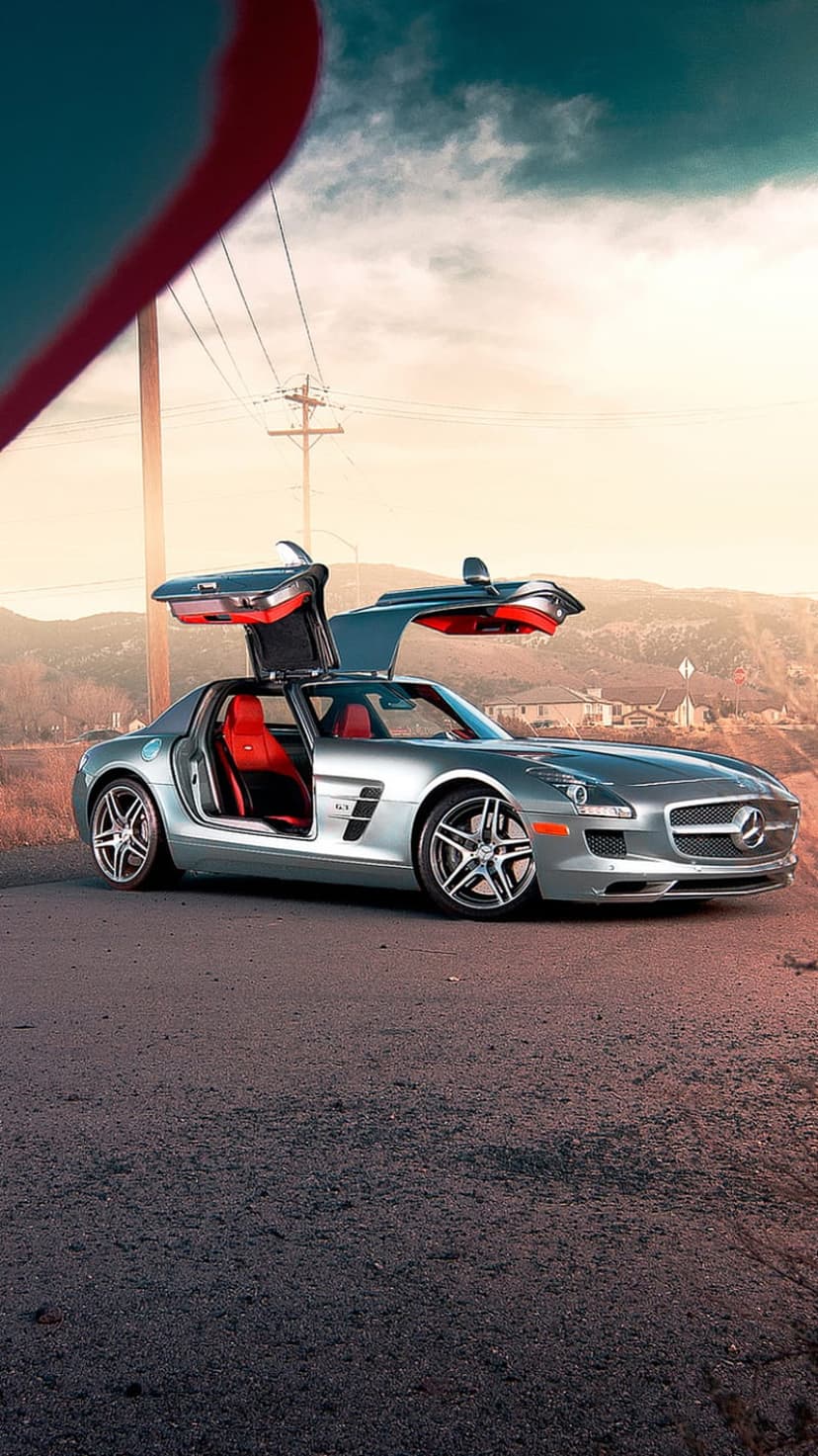 Silver Mercedes SLS AMG with open gullwing doors and red interior parked on a mountain road during a hazy sunset.