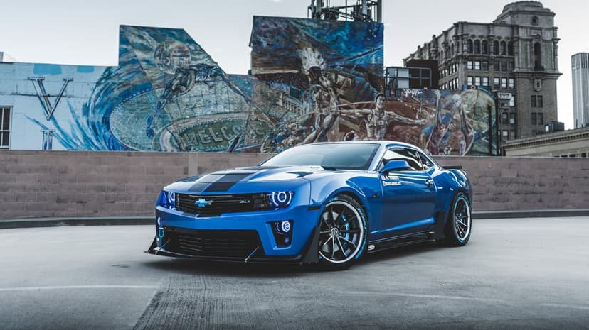 A blue Chevrolet Camaro ZL1 with halo headlights parked on a rooftop in front of a large colorful mural.