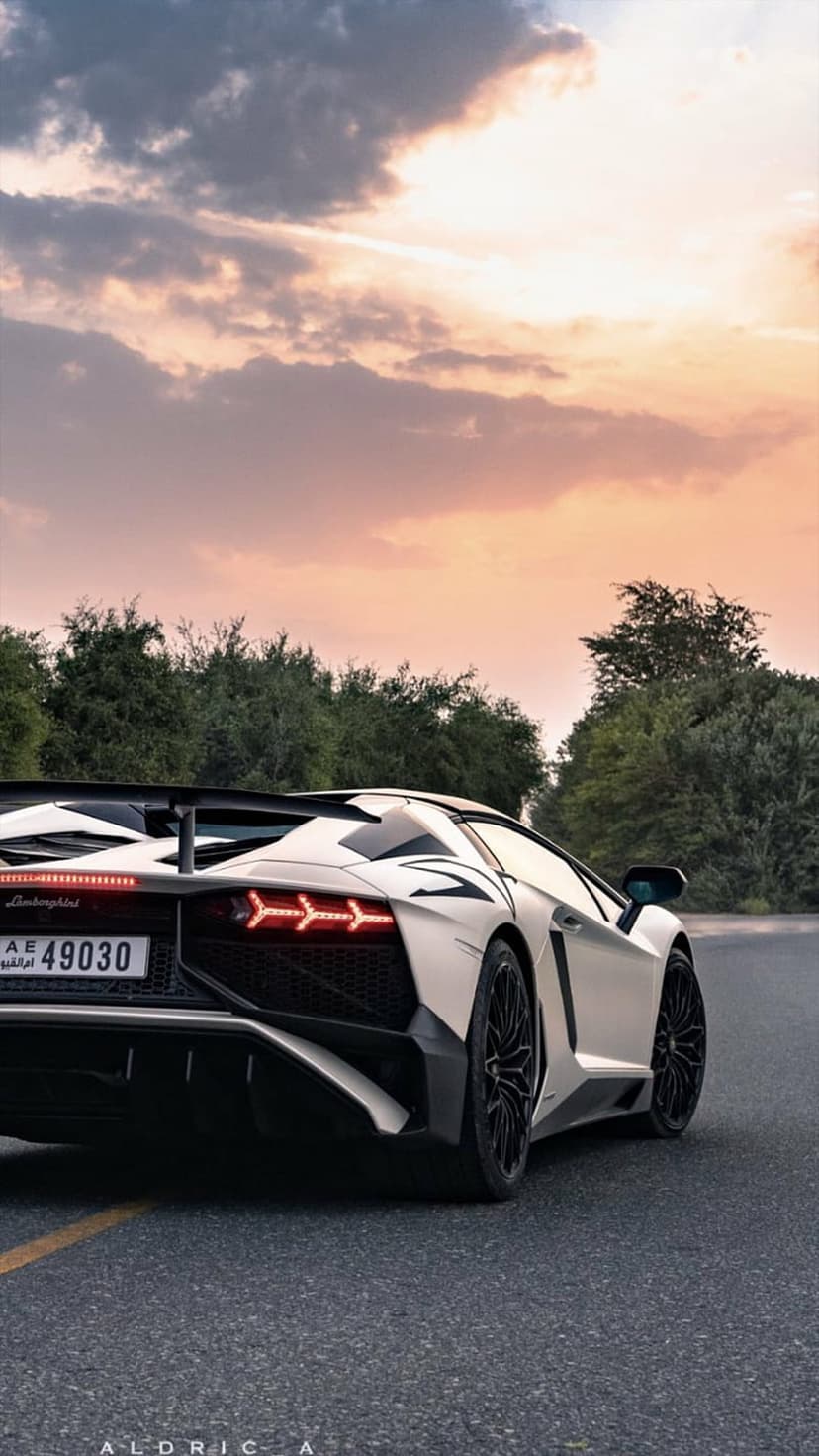 White Lamborghini Aventador rear with glowing LED tail lights parked against a dramatic orange sunset sky.