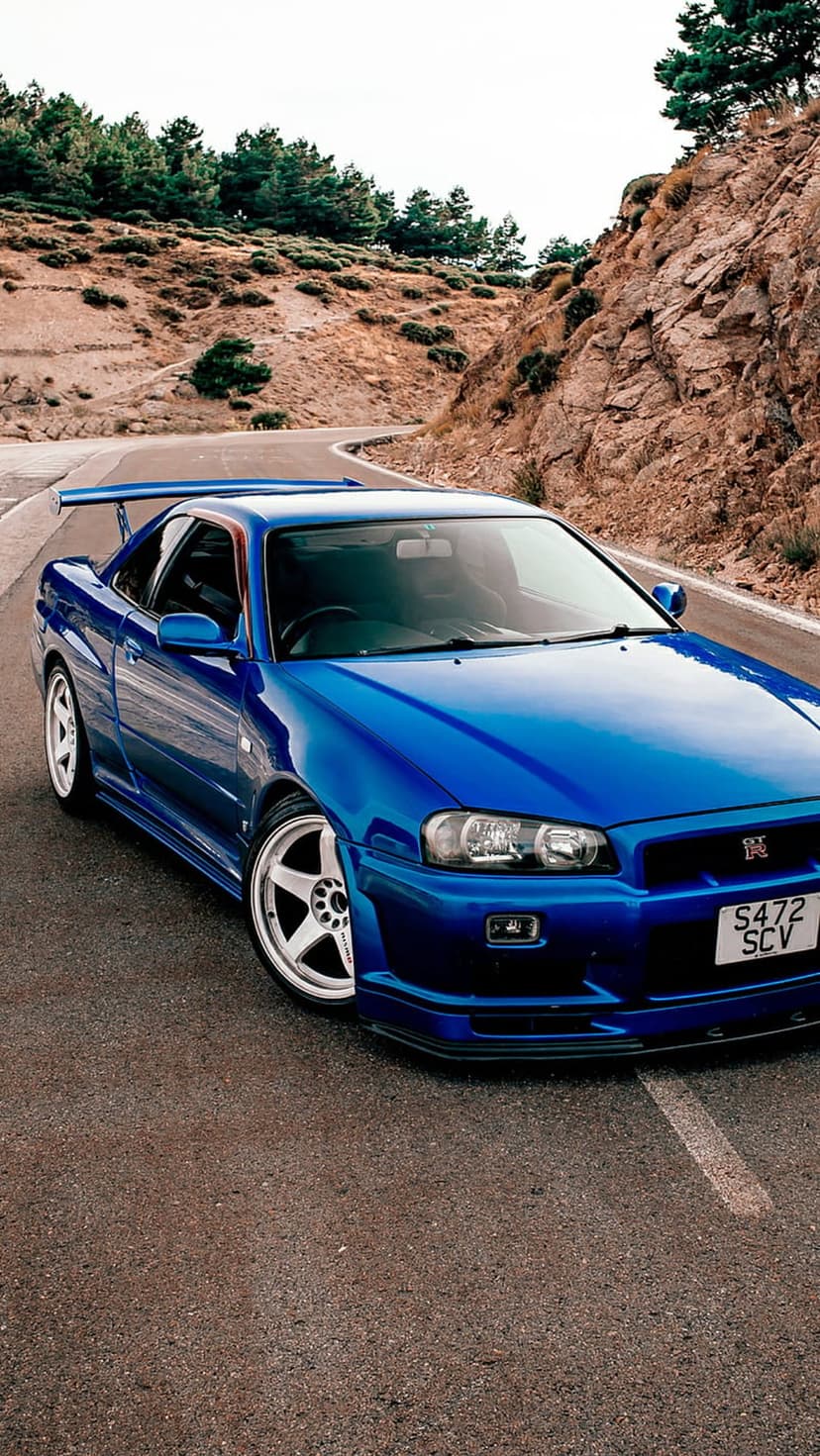 Blue Nissan Skyline GT-R R34 sports car driving on a winding mountain road with green trees and rocky cliffs.