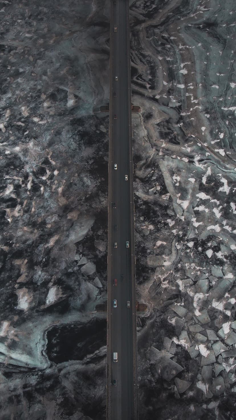 A narrow bridge crosses deep blue water covered in fractured white ice sheets from an aerial perspective.