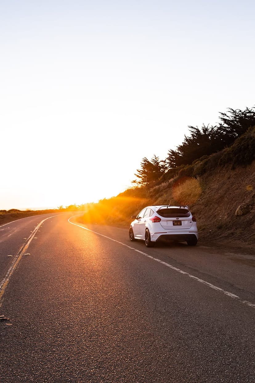 White car driving on a winding coastal road with a bright orange sunset over the ocean and green hills.