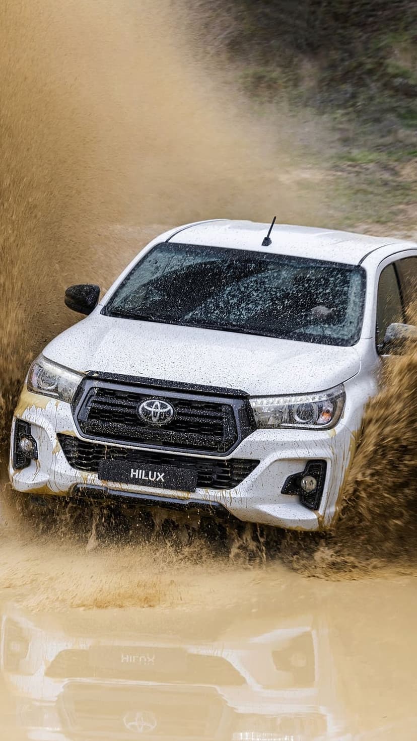 White Toyota Hilux truck splashes through a deep muddy puddle with water spraying from the rugged front tires.