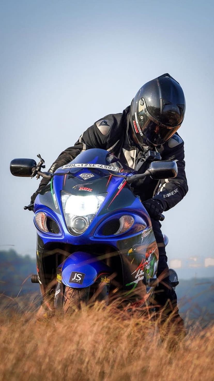 Motorcyclist in black gear on a blue Suzuki Hayabusa leaning forward in tall dry grass under a hazy sky.