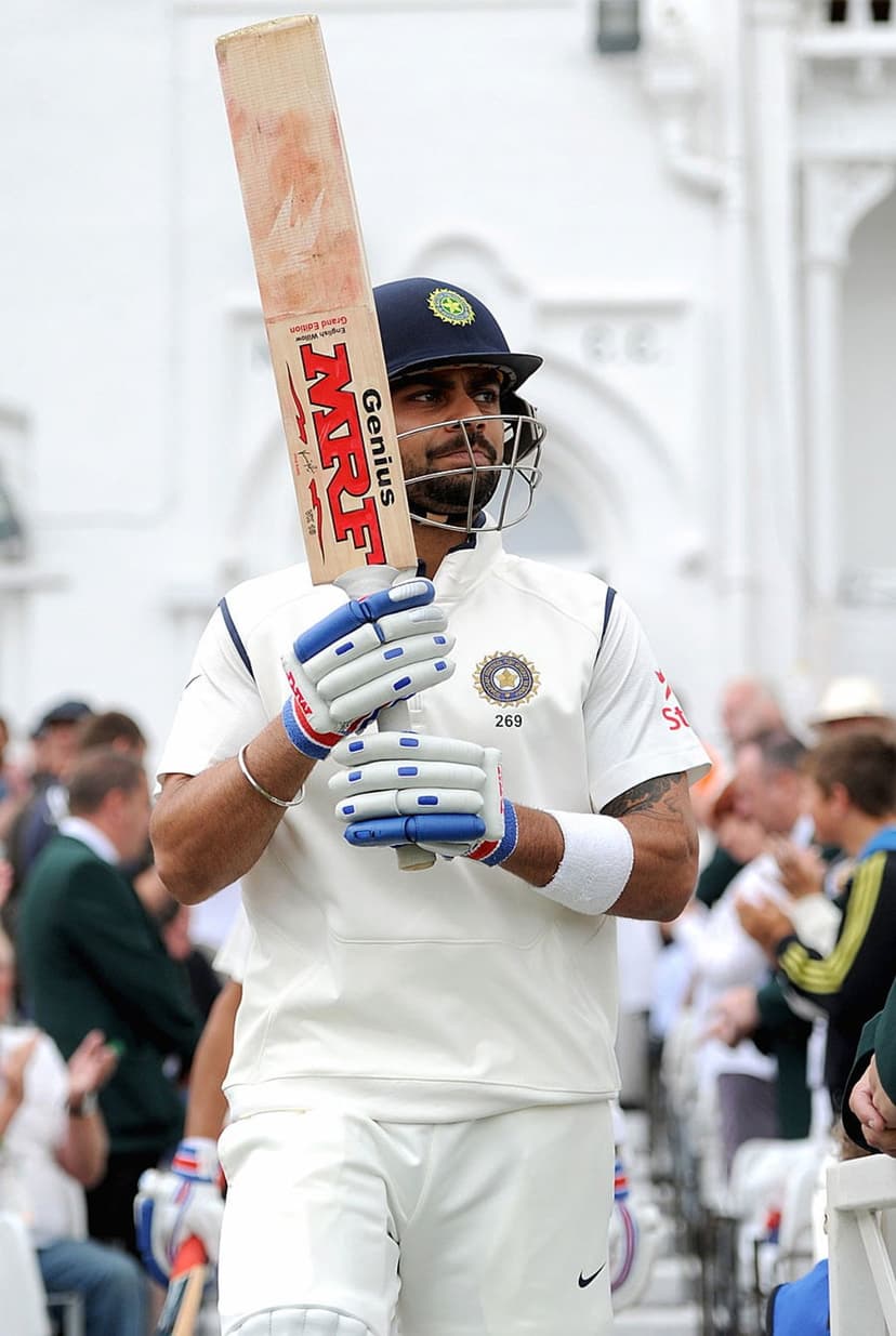 Virat Kohli in white cricket gear walks on a stadium field holding a bat and wearing a protective helmet.