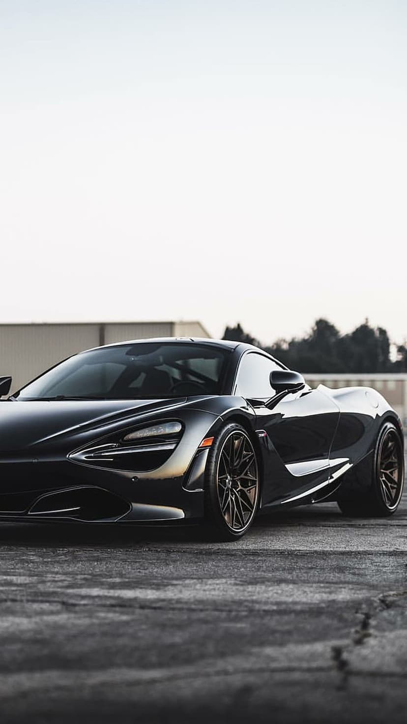 Black McLaren 720S supercar parked on gray asphalt with a low-angle view and industrial buildings in background.