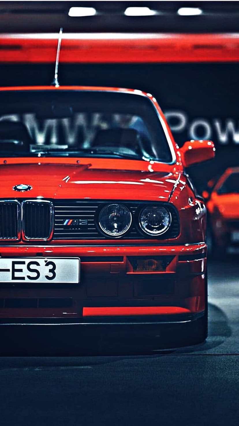 Red BMW M3 E30 front view featuring dual round headlights, black kidney grille, and vibrant paint in a garage.