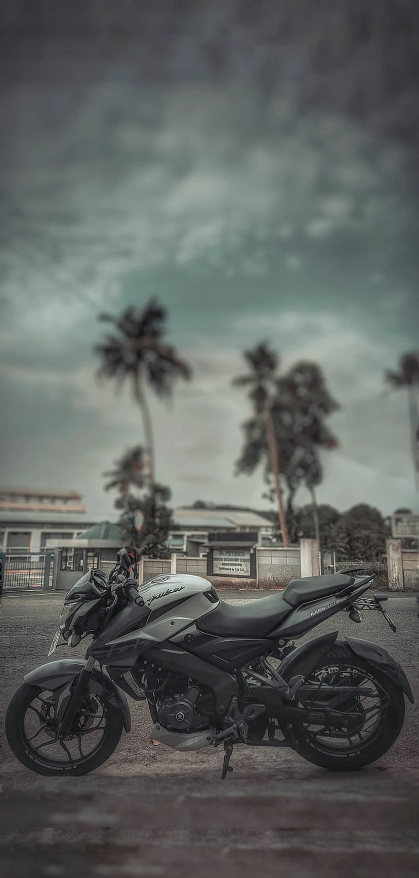 Black and white motorcycle parked on pavement with blurred palm trees and a heavy overcast sky in the background.