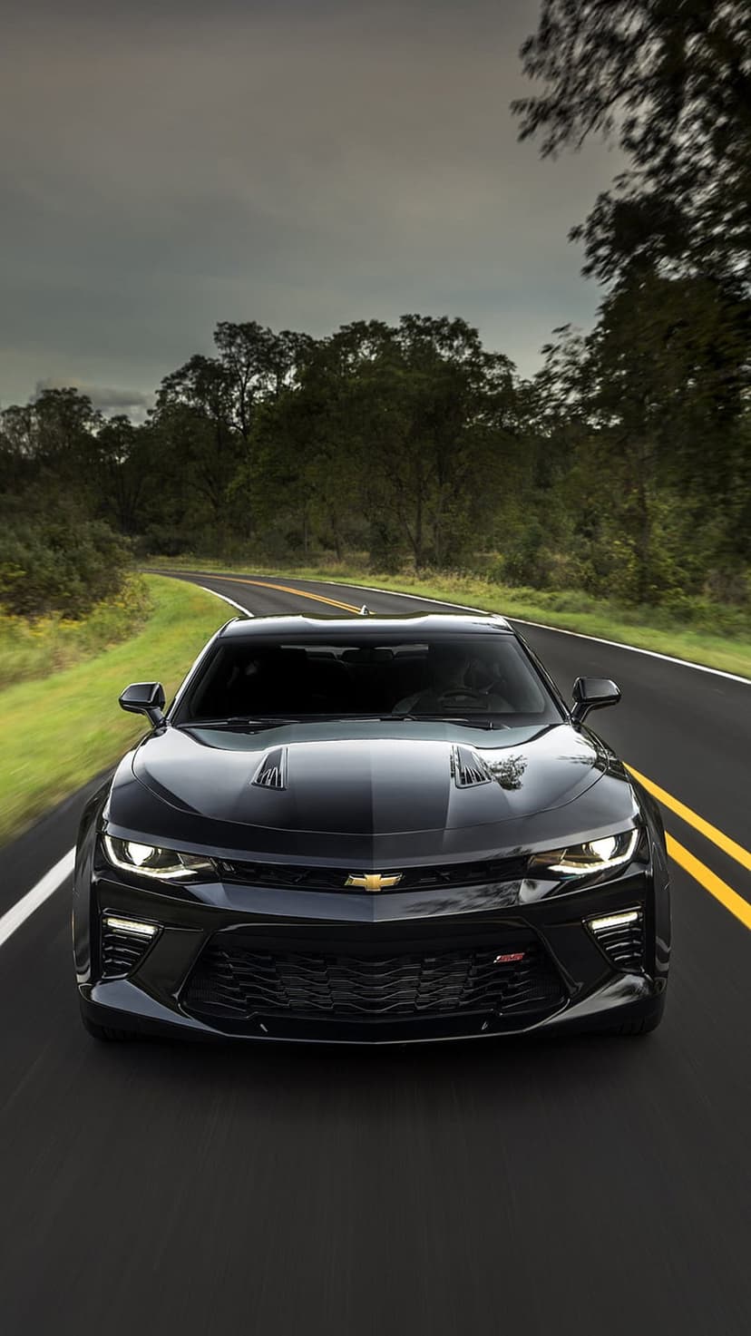 Black Chevrolet Camaro SS with lit headlights driving on a curved asphalt road through a dense green forest.