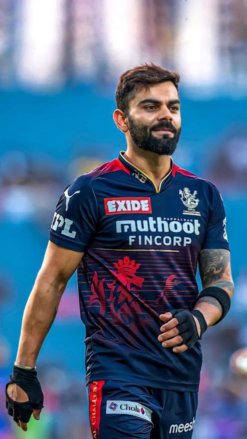 Virat Kohli in a navy and red RCB jersey stands with a focused gaze against a blurred stadium background.