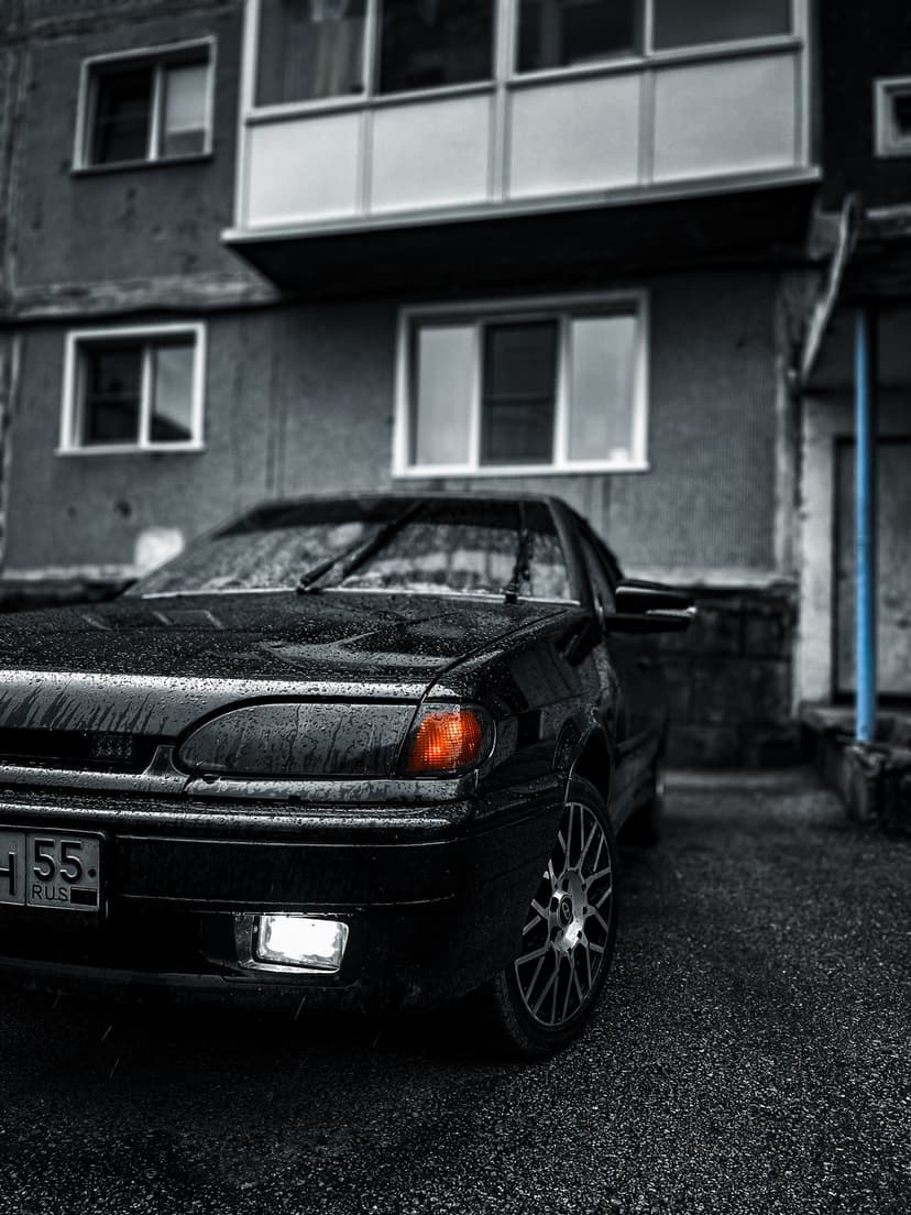 Black sedan with glowing headlights parked on wet asphalt near a grey apartment building on a rainy urban night.