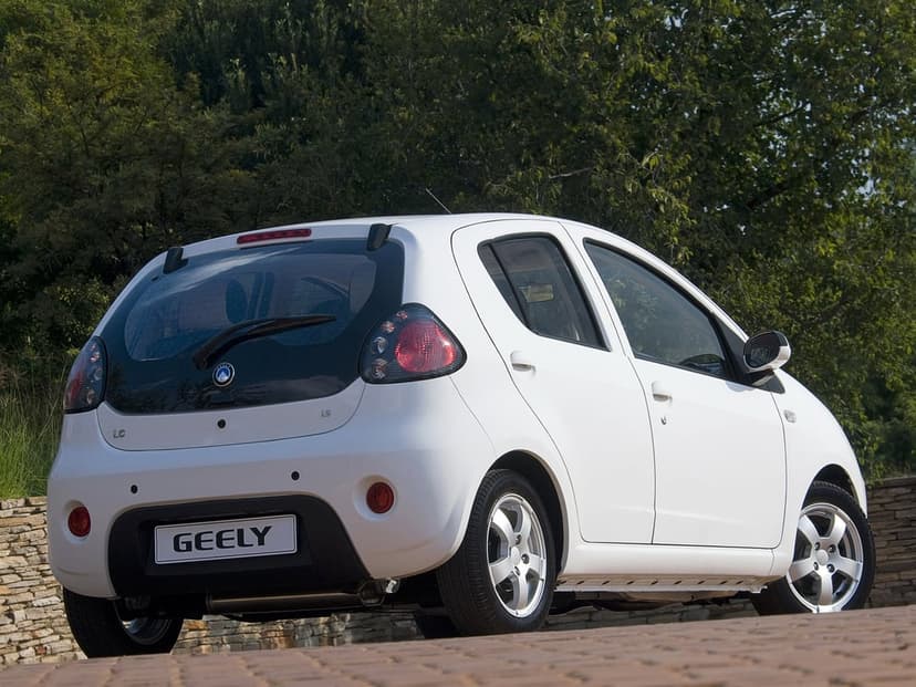 White Geely LC hatchback parked on a brick road with lush green trees in the background under daylight.