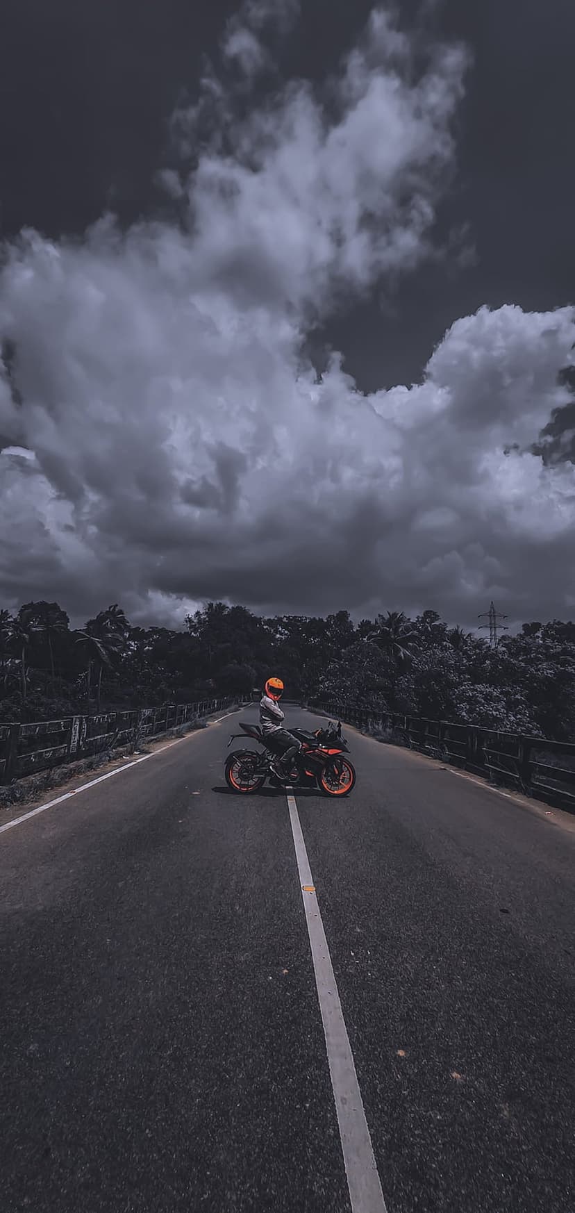 Motorcyclist in a yellow helmet on an orange bike pauses on a bridge road under a dark, dramatic stormy sky.