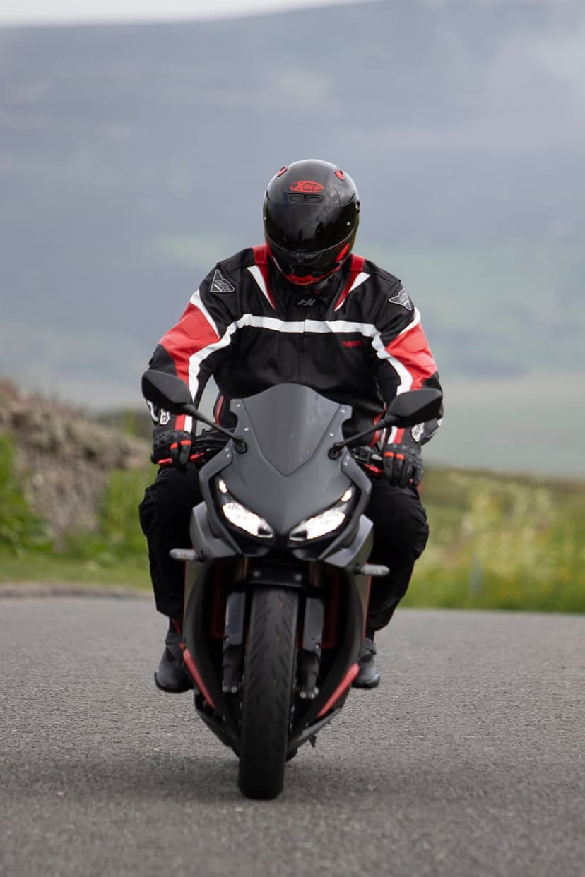 Motorcyclist in red and black gear leans a sportbike on a winding asphalt road with blurred hills in the distance.