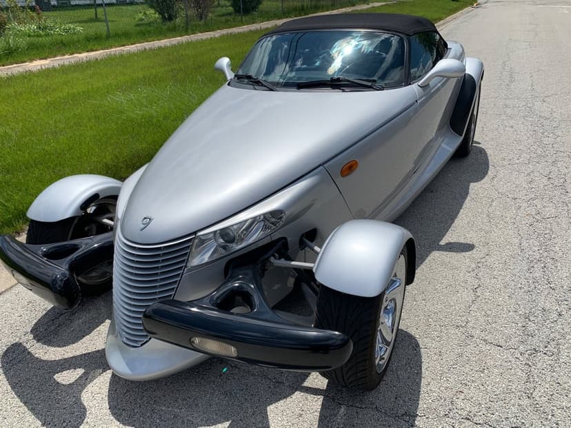 Silver Plymouth Prowler convertible with open-wheel front fenders parked on an asphalt road in bright sunlight.