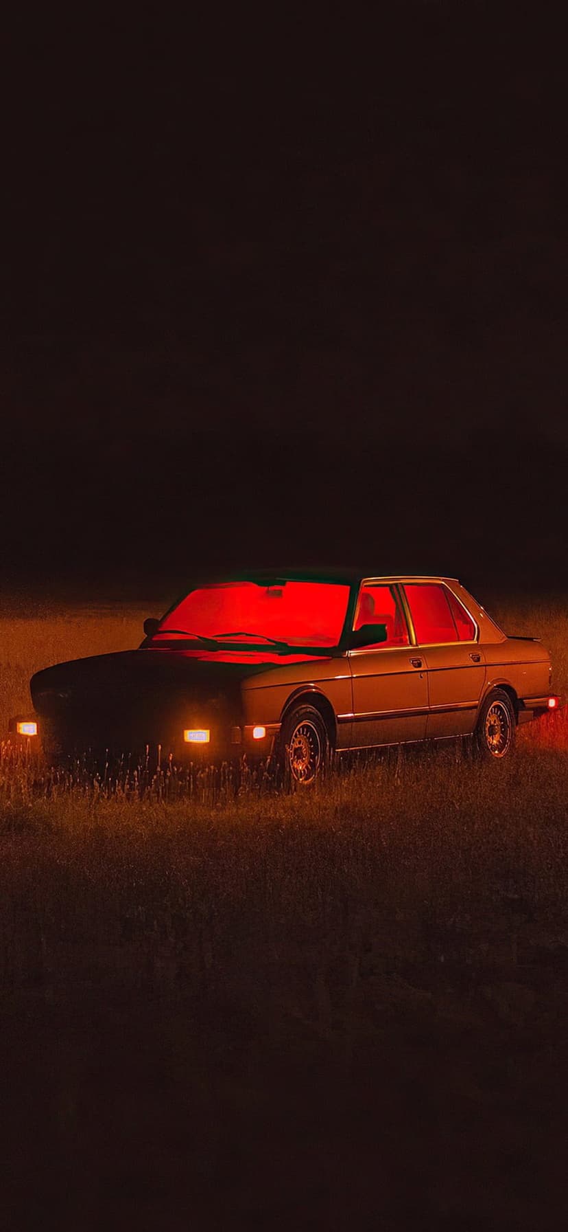 Vintage car with glowing red headlights parked in a dark grassy field at night under a moody atmosphere.