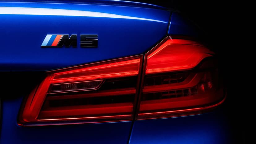 Blue BMW M5 rear trunk with a chrome badge and glowing red LED taillight against a dark background.