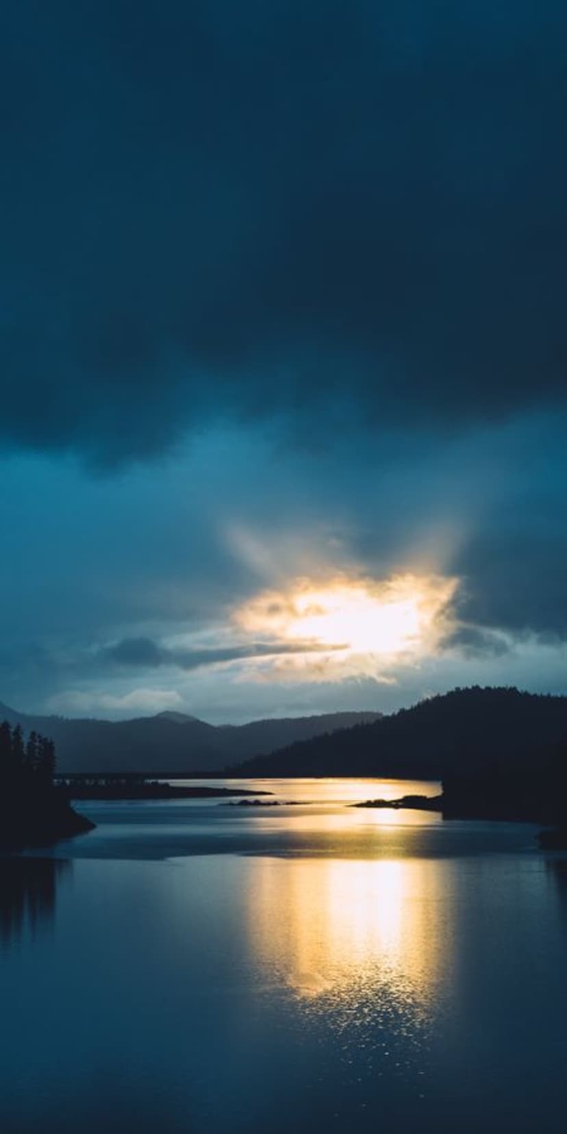 Golden sunlight piercing through clouds reflects on a calm lake surface between dark silhouetted mountains.