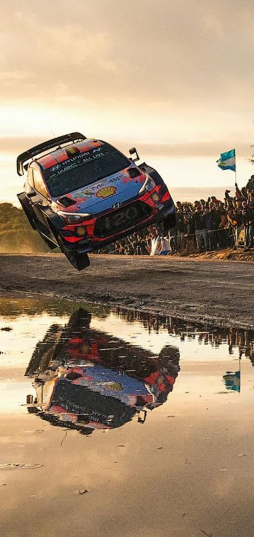 Hyundai rally car leaping through the air with a clear reflection in a puddle during an outdoor race event.