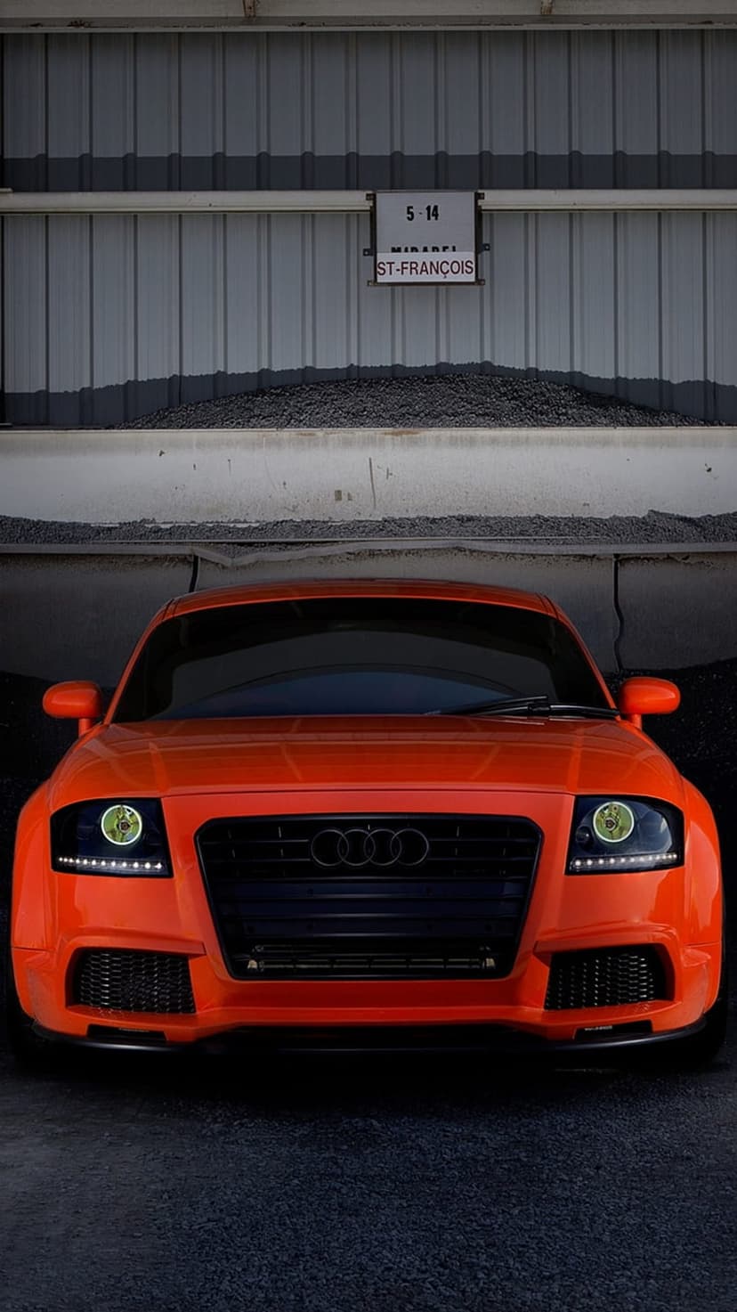 Orange Audi TT front view parked against a corrugated metal wall in an industrial setting with a street sign.