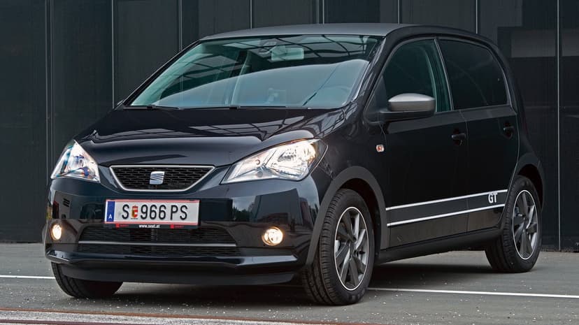 Black SEAT Mii GT city car with glowing headlights parked in front of a dark textured industrial wall.