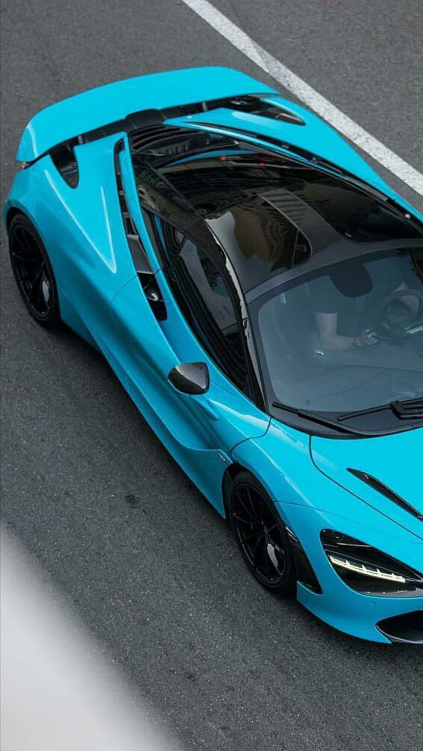 Blue McLaren 720S supercar captured from a top-down perspective while driving on a textured grey asphalt road.