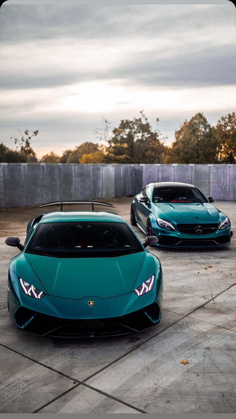 Teal Lamborghini Huracan and Mercedes-AMG C63 parked on concrete near autumn trees under a moody cloudy sky.