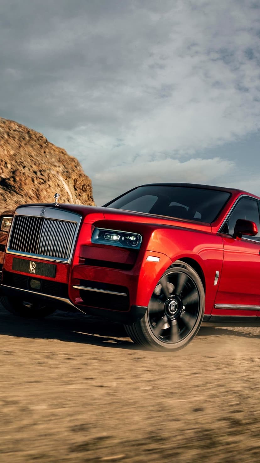 Red luxury SUV driving on a dirt road against rocky hills and a cloudy sky with motion-blurred wheels.