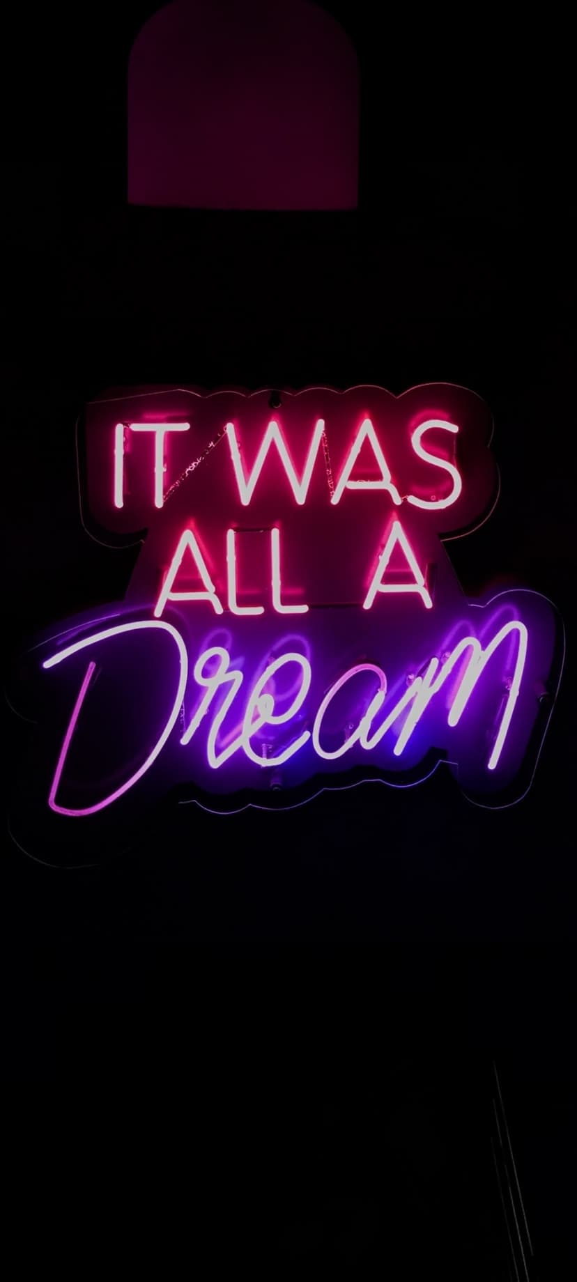 Neon sign reading "It Was All A Dream" glows in hot pink and purple hues against a dark, shadowy background.