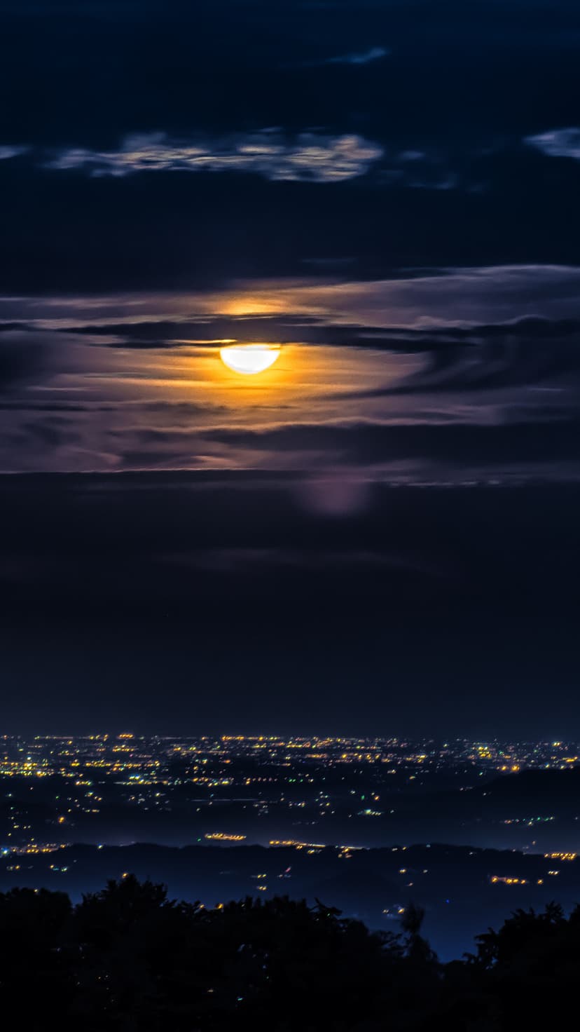 Full moon glowing through dark clouds above a distant city skyline with shimmering lights at night.